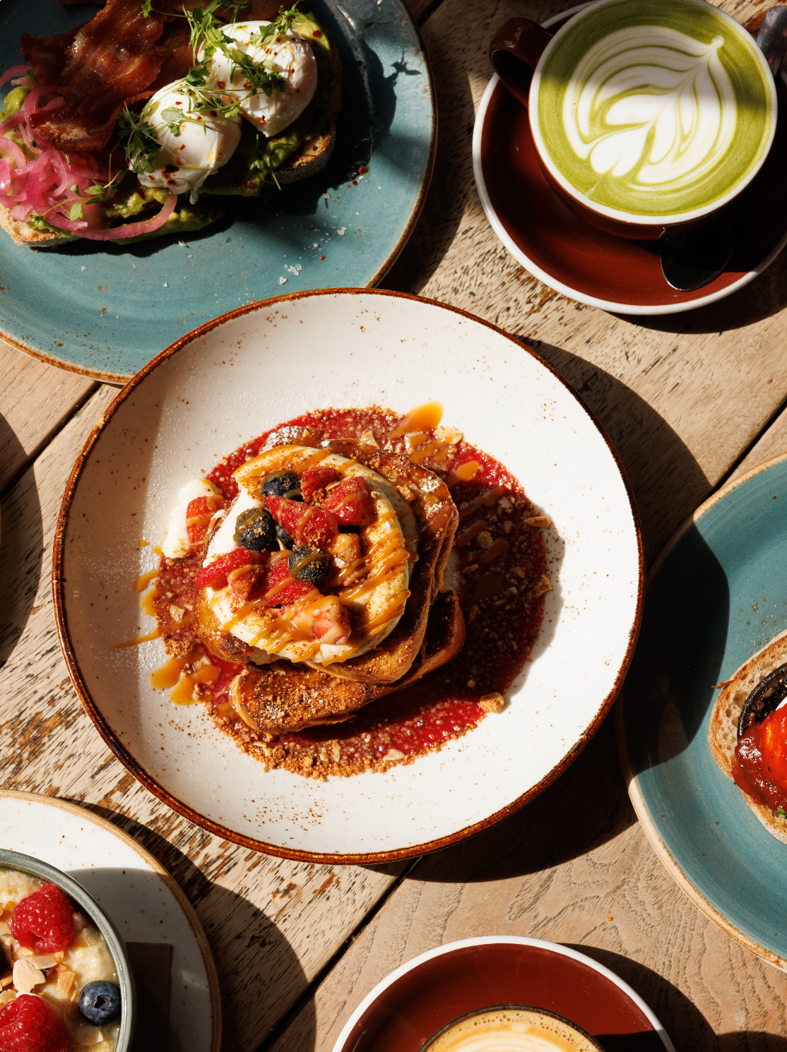 Overhead view of a plated breakfast dish on a rustic wooden table, surrounded by other food and a cup of latte art coffee. The main plate features Federal French Toast.