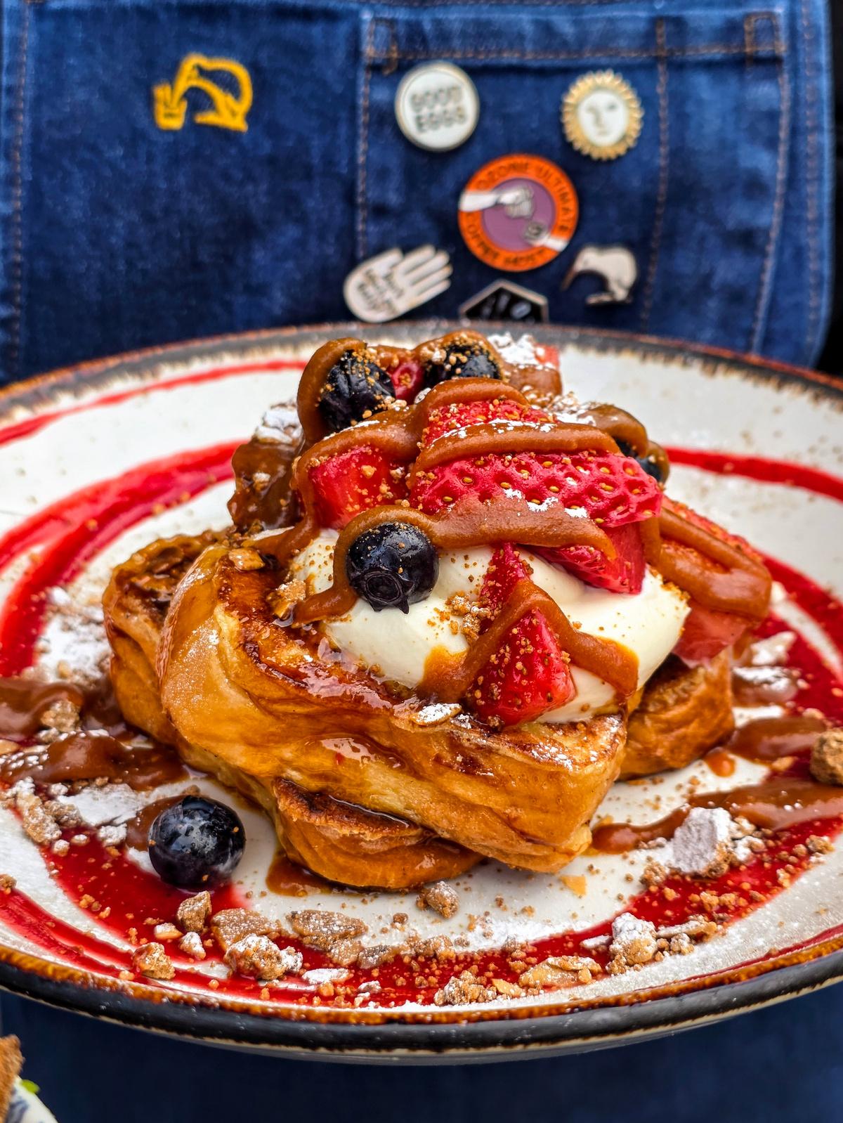 A plated Federal French toast topped with vanilla mascarpone, strawberries, blueberries, powdered sugar, and drizzles of salted caramel sauce and raspberry coulee, set against a denim apron background with pins.