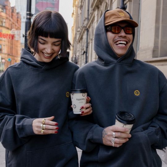Two people wearing dark hoodies and sunglasses, holding coffee cups on a city street.