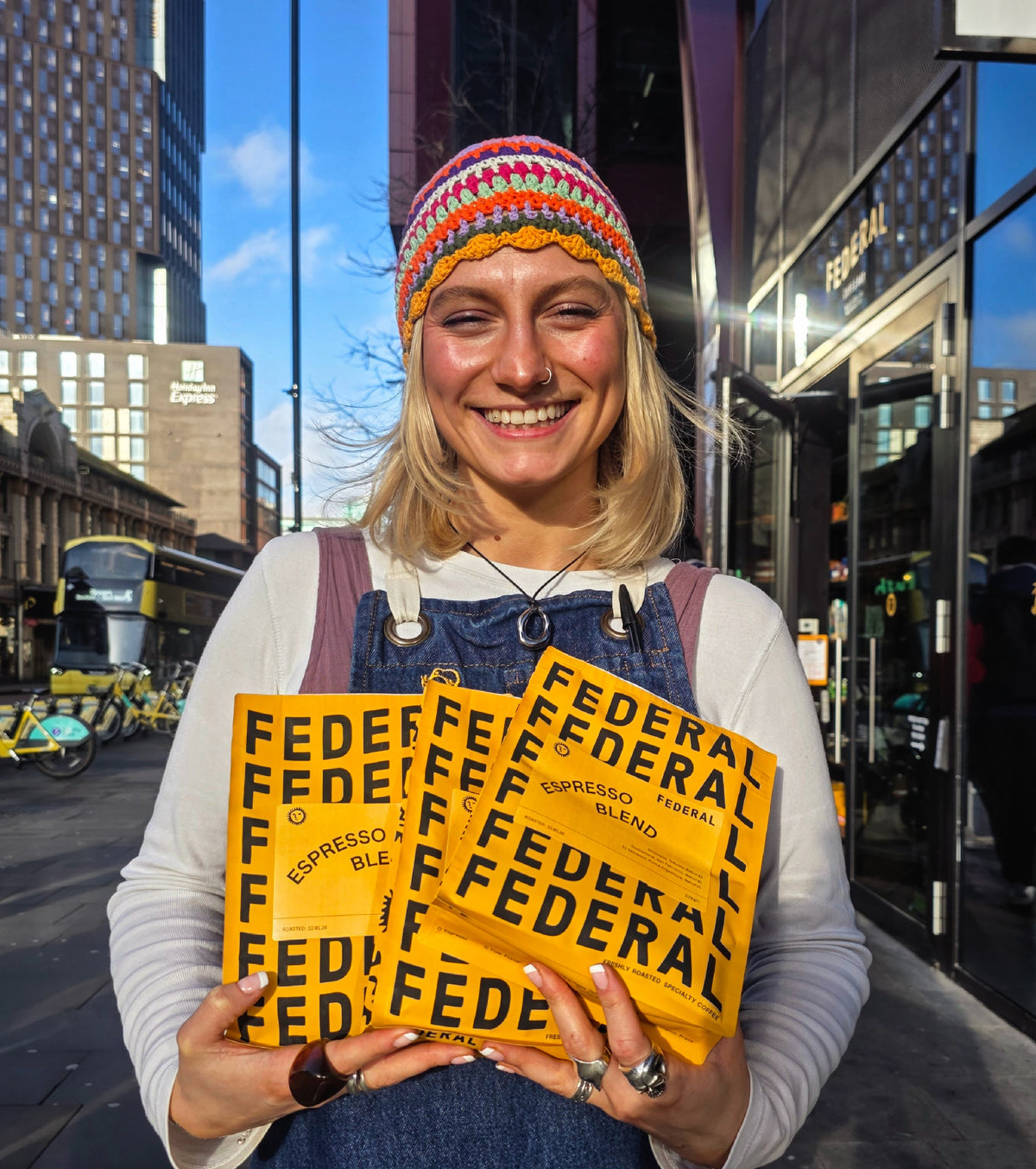 A smiling woman outdoors in an urban setting holds three bright yellow coffee bags with bold black text reading 'FEDERAL'. She wears a multicolored knit beanie, a white long-sleeve top, and blue denim apron.