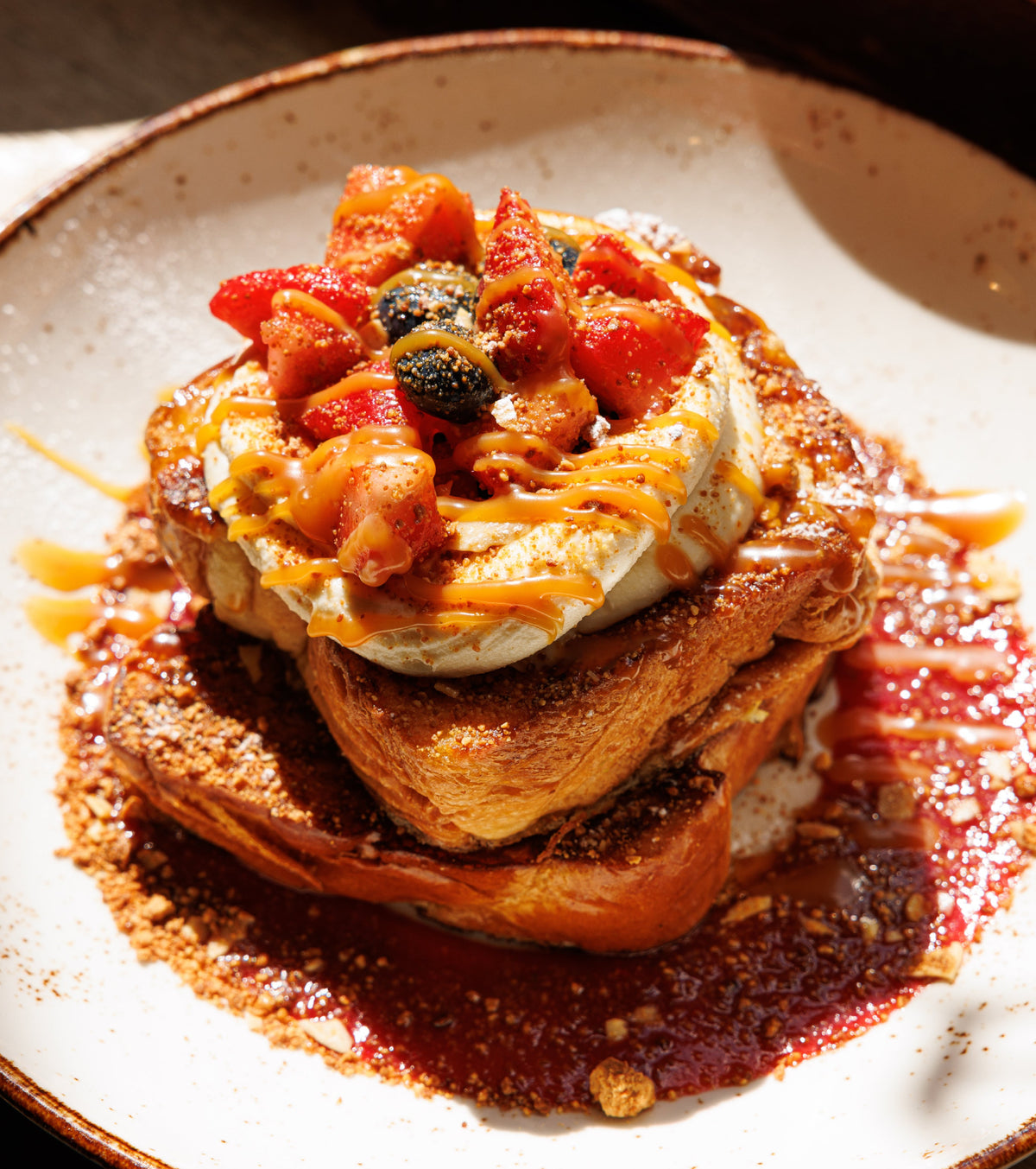 A plated French toast featuring a stack of golden-brown brioche topped with vanilla mascarpone, fresh berries and caramel drizzle on a white plate with a brown rim.