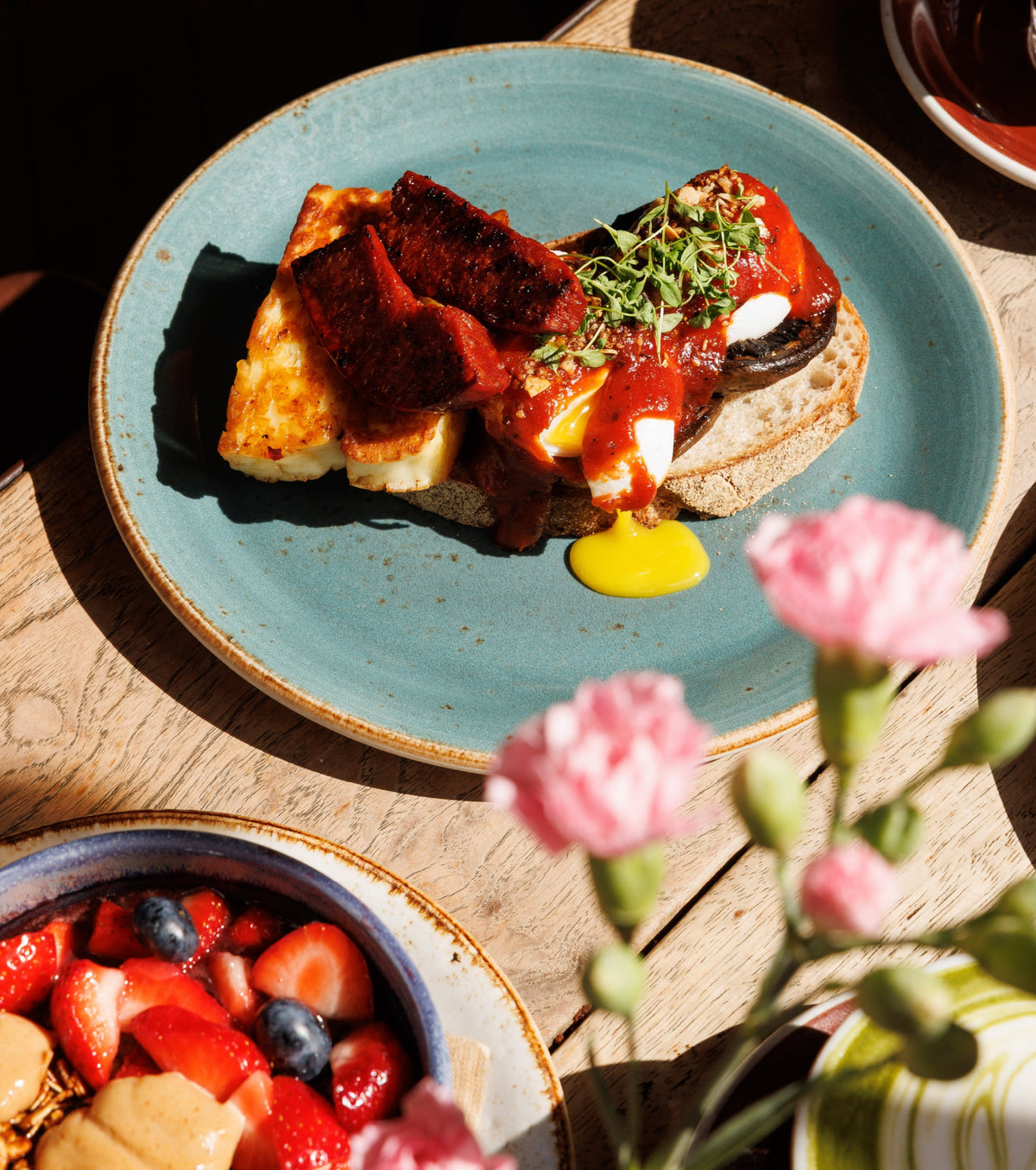 A plated breakfast dish on a rustic wooden table, featuring poached eggs, halloumi, mushrooms, chorizo served on a blue-green ceramic plate with pink flowers in the foreground.